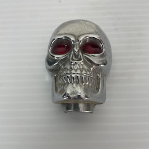 May include: A chrome skull-shaped ornament with red eye sockets. The skull has detailed facial features, including a nose and teeth. The object is likely a decorative item or part of a larger assembly.
