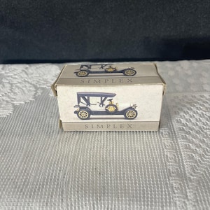 May include: A small, beige box featuring a vintage car illustration. The car is black with gold wheels and accents, and the word "SIMPLEX" is printed above and below the image. The box is on a white, textured surface.