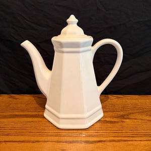 May include: A white ceramic mug with a black handle and the text "But First, Coffee" in a black, handwritten-style font. The mug is on a white surface.