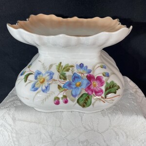 May include: White ceramic pedestal bowl with a scalloped rim and a flared base. The bowl is decorated with blue and pink floral designs and green leaves. The rim has a gold-colored trim.
