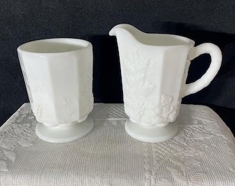 May include: A white milk glass creamer and sugar bowl set. The creamer has a handle and spout, while the sugar bowl is a small, open cup. Both pieces have a pedestal base and a grape and leaf design.