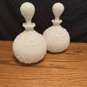 May include: Two white, vintage perfume bottles with ornate, raised floral designs. Each bottle has a round body and a decorative stopper. The bottles are displayed on a wooden surface against a dark background.