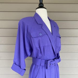 May include: A vibrant purple jumpsuit with a tailored blazer-style top and a matching belt. The jumpsuit features a collared neckline, two front pockets, and three-quarter sleeves. The belt cinches the waist, and the fabric appears to be a lightweight linen or similar material.