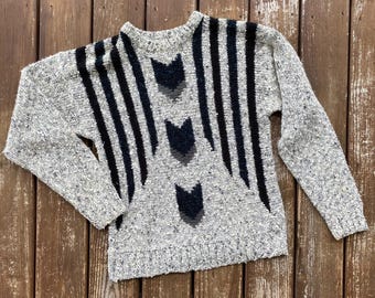 Può includere: Un maglione grigio a maglia con strisce verticali nere e disegni a forma di freccia grigio scuro. Il maglione ha maniche lunghe e scollatura e orlo a coste. Il tessuto ha una trama macchiettata.