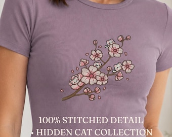 Pode incluir: Um top curto de manga curta em tecido canelado malva com um bordado de um ramo de cerejeira em flor rosa com pequenos gatos brancos escondidos entre as flores. O texto 100% STITCHED DETAIL HIDDEN CAT COLLECTION está impresso em branco.