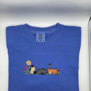K&ouml;nnte beinhalten: Ein blaues T-Shirt mit einer Stickerei von drei Katzen, die in einem Feld mit Blumen und einem Schmetterling sitzen. Die Katzen sind schwarz-wei&szlig;, grau und orange. Das T-Shirt hat ein Comfort Colors Markenetikett am Kragen mit Gr&ouml;&szlig;e M.