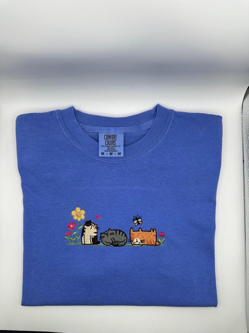 K&ouml;nnte beinhalten: Ein blaues T-Shirt mit einer Stickerei von drei Katzen, die in einem Feld mit Blumen und einem Schmetterling sitzen. Die Katzen sind schwarz-wei&szlig;, grau und orange. Das T-Shirt hat ein Comfort Colors Markenetikett am Kragen mit Gr&ouml;&szlig;e M.