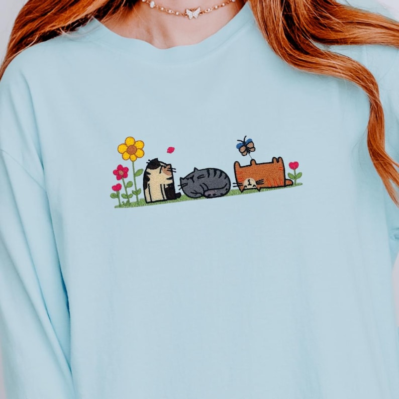 K&ouml;nnte beinhalten: Ein hellblaues Langarmshirt mit einer Stickerei von drei Katzen, die auf einer gr&uuml;nen Grasfl&auml;che mit bunten Blumen und einem Schmetterling ruhen. Die Katzen umfassen eine schwarz-wei&szlig;e Katze, eine graue getigerte Katze und eine orangefarbene Katze.