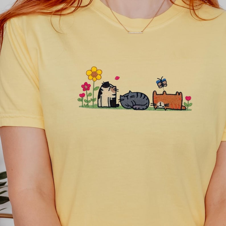 K&ouml;nnte beinhalten: Ein hellgelbes T-Shirt mit einer Stickerei von drei Katzen in einem Garten. Die Katzen sind in Schwarz-Wei&szlig;, Grau und Orange dargestellt, umgeben von bunten Blumen und einem kleinen blauen Schmetterling &uuml;ber der orangefarbenen Katze.