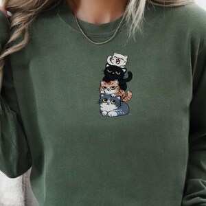 以下が含まれることがあります： 4匹の漫画の猫が積み重なった刺繍デザインが施されたグリーンのスウェットシャツ。猫は白、黒、オレンジ、グレーです。スウェットシャツはクルーネックと長袖です。