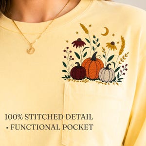 May include: A pale yellow long-sleeve shirt featuring a functional chest pocket and an embroidered design of three pumpkins, wheat stalks, flowers, and a crescent moon. The text 100% STITCHED DETAIL • FUNCTIONAL POCKET is printed on the shirt.