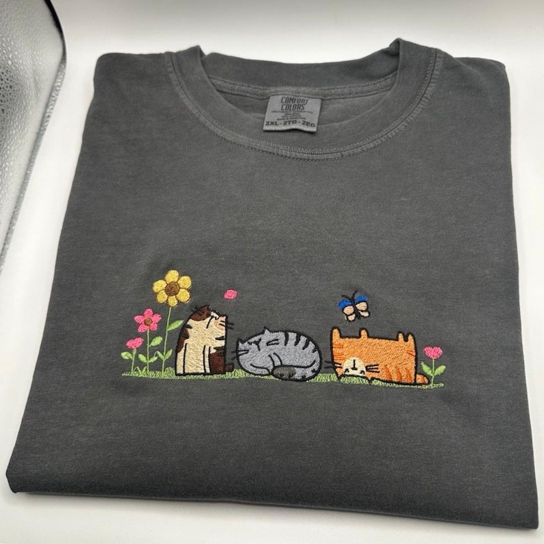 K&ouml;nnte beinhalten: Ein anthrazitfarbenes T-Shirt mit einer Stickerei von drei Katzen, die in einem Garten ruhen. Das Motiv zeigt eine braune getigerte Katze, eine graue getigerte Katze und eine orangefarbene Katze, umgeben von bunten Blumen, einem rosa Bl&uuml;tenblatt und einem blauen Schmetterling.