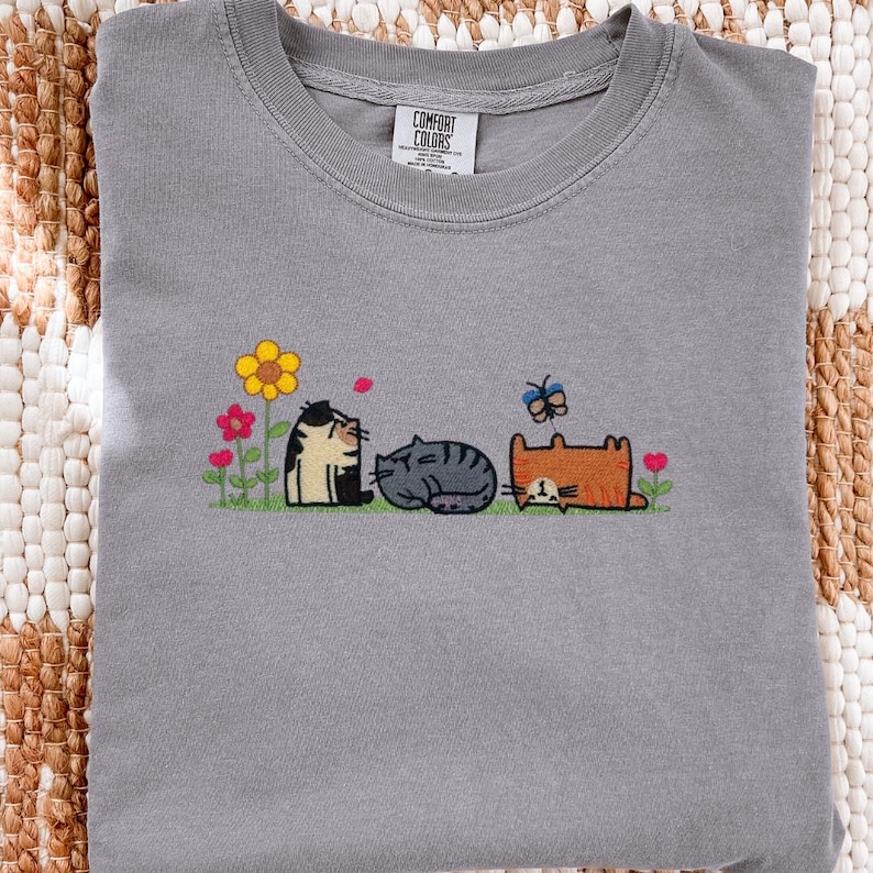 K&ouml;nnte beinhalten: Ein graues T-Shirt mit Rundhalsausschnitt und einer Stickerei von drei Katzen in einem Garten. Die Katzen sind schwarz-wei&szlig;, grau gestreift und orange. Das Motiv zeigt eine gelbe Blume, rosa Blumen, gr&uuml;ne St&auml;ngel und einen blauen Schmetterling.