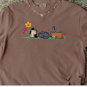 K&ouml;nnte beinhalten: Ein braunes T-Shirt mit einer Stickerei von drei Katzen, die auf gr&uuml;nem Gras sitzen, umgeben von bunten Blumen und einem Schmetterling. Die Katzen sind schwarz-wei&szlig;, grau getigert und orange. Dazu wird eine zarte Perlenkette getragen.