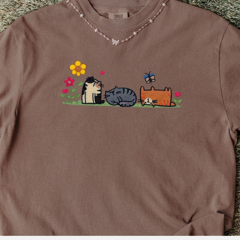 K&ouml;nnte beinhalten: Ein braunes T-Shirt mit einer Stickerei von drei Katzen, die auf gr&uuml;nem Gras sitzen, umgeben von bunten Blumen und einem Schmetterling. Die Katzen sind schwarz-wei&szlig;, grau getigert und orange. Dazu wird eine zarte Perlenkette getragen.