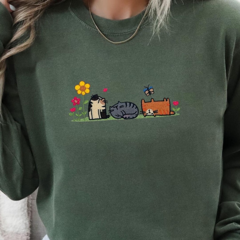K&ouml;nnte beinhalten: Ein olivgr&uuml;nes Sweatshirt mit Rundhalsausschnitt und einer Stickerei von drei Katzen auf einer Wiese. Die Katzen sind schwarz-wei&szlig;, grau und orange. Das Motiv zeigt eine gelbe Sonnenblume, rosa Blumen und einen blauen Schmetterling dar&uuml;ber.