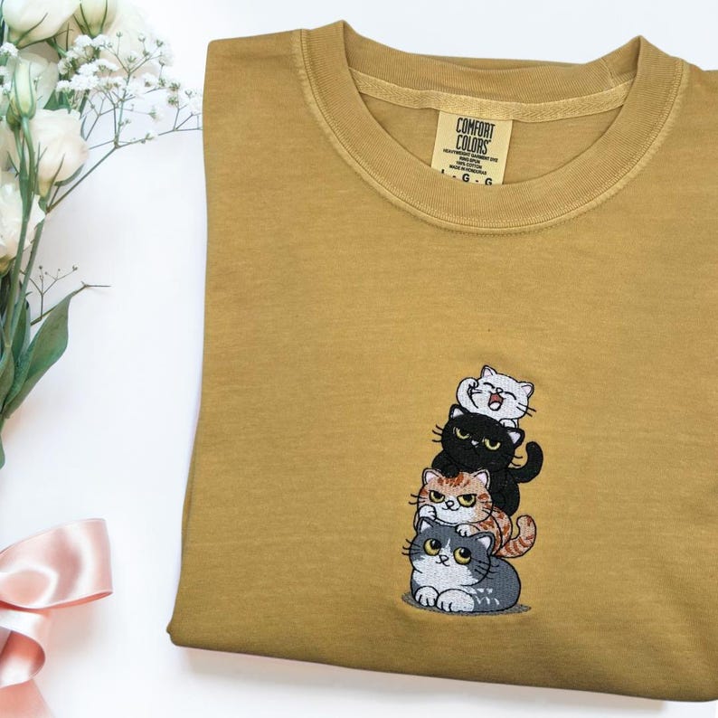 以下が含まれることがあります： マスタード色のTシャツ。4匹の漫画の猫が積み重なった刺繍デザインが施されています。猫は黒、グレー、オレンジ、白です。シャツはクルーネックで、「Comfort Colors」のタグが付いています。
