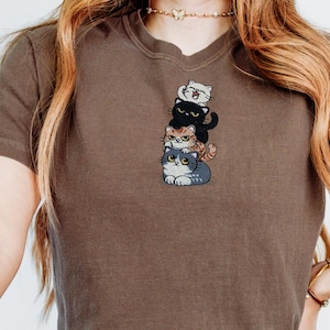 以下が含まれることがあります： 茶色のTシャツ。4匹の漫画の猫が積み重なった刺繍デザイン。猫は白、黒、オレンジのトラ柄、グレーと様々な色。Tシャツは無地の茶色です。