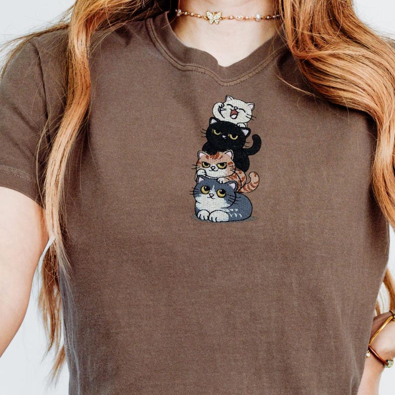 以下が含まれることがあります： 茶色のTシャツ。4匹の漫画の猫が積み重なった刺繍デザイン。猫は白、黒、オレンジのトラ柄、グレーと様々な色。Tシャツは無地の茶色です。