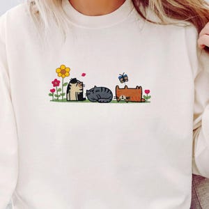 K&ouml;nnte beinhalten: Ein cremefarbenes Sweatshirt mit Rundhalsausschnitt und einer Stickerei von drei Katzen auf einer Wiese. Das Motiv zeigt eine gelbe Blume, rote Blumen, einen blauen Schmetterling sowie drei Katzen: eine Gl&uuml;ckskatze, eine graue Tabbykatze und eine orangefarbene Katze.
