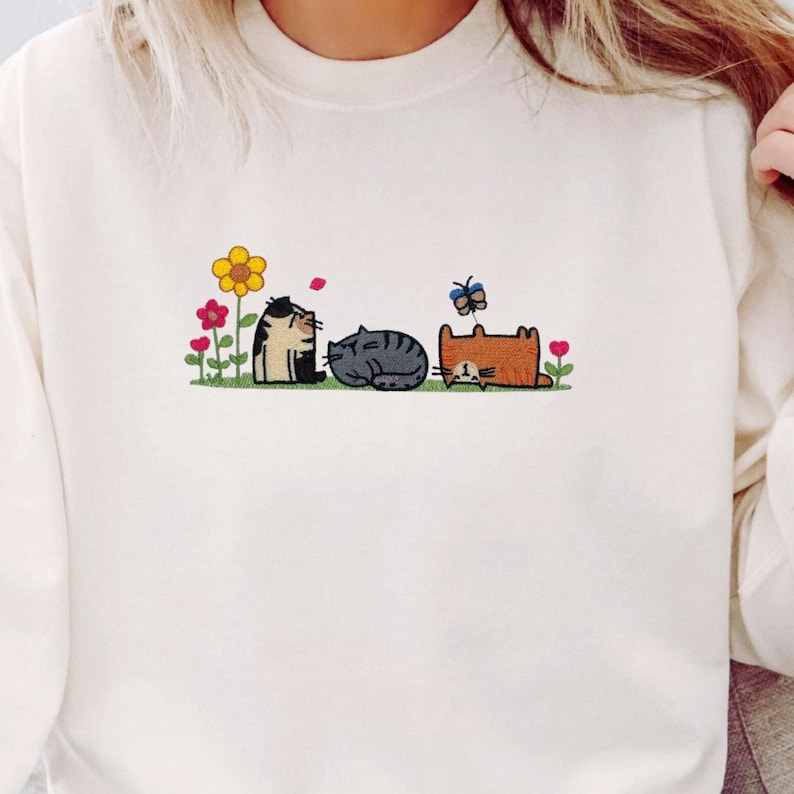 K&ouml;nnte beinhalten: Ein cremefarbenes Sweatshirt mit Rundhalsausschnitt und einer Stickerei von drei Katzen auf einer Wiese. Das Motiv zeigt eine gelbe Blume, rote Blumen, einen blauen Schmetterling sowie drei Katzen: eine Gl&uuml;ckskatze, eine graue Tabbykatze und eine orangefarbene Katze.