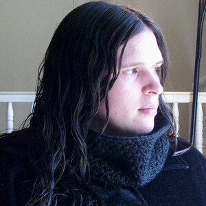 Könnte beinhalten: Seitenprofil einer Person mit langen, dunklen, nassen Haaren. Sie trägt einen dunkelgrauen, gestrickten Rundschal und einen schwarzen Mantel. Der Hintergrund ist neutral.