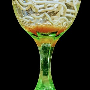 May include: A green-tinted glass with a clear liquid containing numerous white, worm-like objects. The glass has a green stem and base, set against a black background.