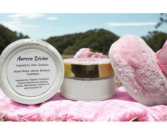 May include: Two white jars of Aurora Divine powder with gold lids and labels, next to a pink fluffy object. The labels list ingredients and scent notes of vanilla, bourbon, and cashmere. The jars sit on a pink surface.