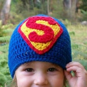 May include: A blue crocheted beanie featuring the Superman logo in red and yellow. The hat is handmade and has a textured appearance. The Superman logo is prominently displayed on the front of the hat.