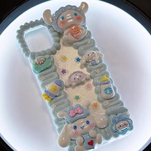 May include: A light blue phone case decorated with various cartoon characters and pastel-colored embellishments. The case features a baby-themed design with stars, a bottle, and a rocket ship. The case has a wavy border.