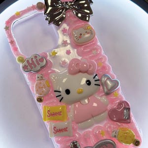 May include: Pink phone case with Hello Kitty design, featuring a 3D bow, hearts, stars, and the word "Sweet". The case has a clear border and is decorated with various charms and embellishments.