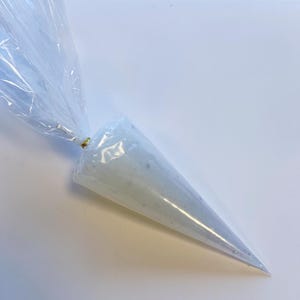 May include: A clear plastic piping bag filled with white frosting. The bag is sealed at the top with a gold twist tie, and the frosting is visible through the transparent material. The bag tapers to a point at the bottom.