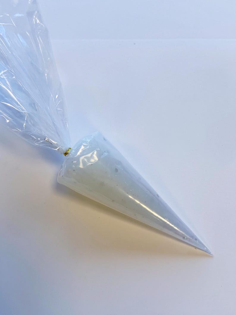 May include: A clear plastic piping bag filled with white frosting. The bag is sealed at the top with a gold twist tie, and the frosting is visible through the transparent material. The bag tapers to a point at the bottom.