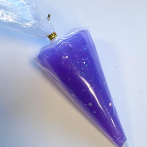 May include: A clear plastic piping bag filled with purple gel and iridescent glitter. The bag is sealed with a gold twist tie. The gel is a vibrant purple color, and the glitter adds a sparkling effect.