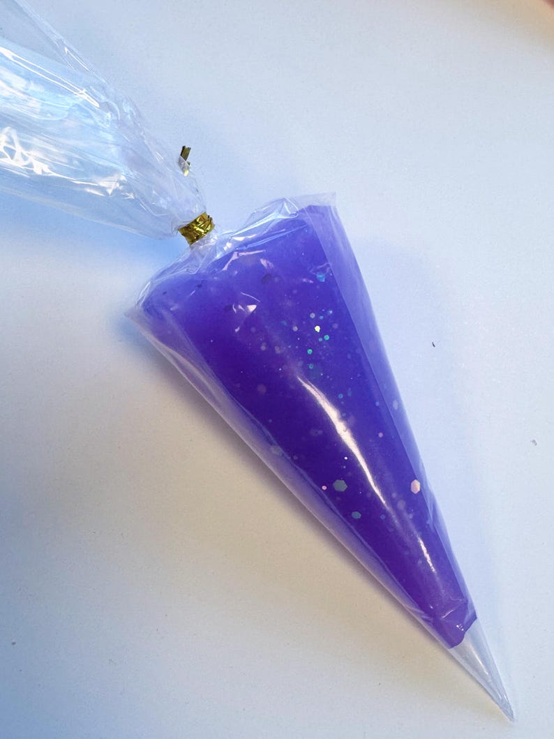 May include: A clear plastic piping bag filled with purple gel and iridescent glitter. The bag is sealed with a gold twist tie. The gel is a vibrant purple color, and the glitter adds a sparkling effect.