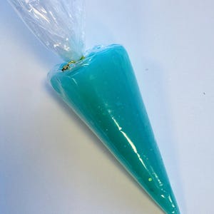 May include: A clear plastic piping bag filled with bright teal frosting. The frosting is a smooth, opaque color with a few flecks of glitter. The bag is sealed at the top with a twist tie, ready for decorating cakes or other baked goods.