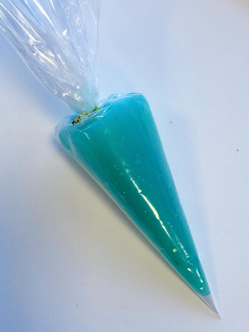 May include: A clear plastic piping bag filled with bright teal frosting. The frosting is a smooth, opaque color with a few flecks of glitter. The bag is sealed at the top with a twist tie, ready for decorating cakes or other baked goods.