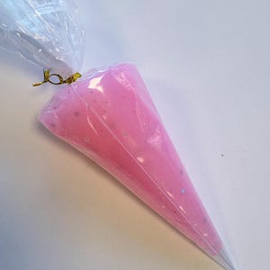 May include: A clear plastic piping bag filled with pink frosting. The frosting has a smooth texture with small, silver-colored specks. The top of the bag is tied with a gold twist tie, and the tip is a silver cone.