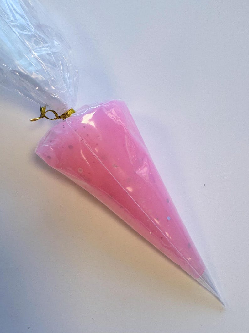 May include: A clear plastic piping bag filled with pink frosting. The frosting has a smooth texture with small, silver-colored specks. The top of the bag is tied with a gold twist tie, and the tip is a silver cone.