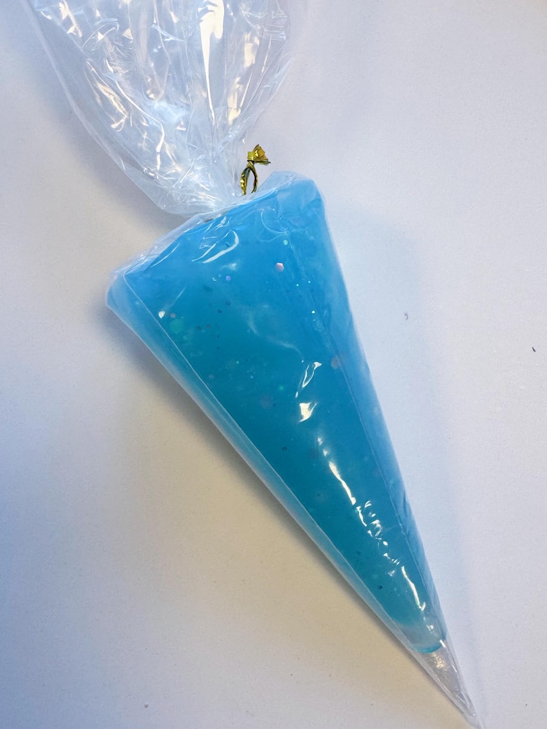 May include: A clear plastic piping bag filled with light blue frosting. The frosting has a slightly glittery appearance. The top of the bag is twisted and secured with a gold-colored twist tie. The bag is on a white surface.
