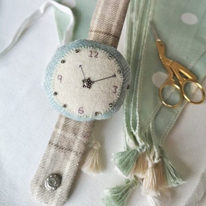 May include: A light blue and brown fabric watch with a white face and black hands. The watch has a decorative button and a tassel on the strap. The watch is surrounded by green fabric with white polka dots.
