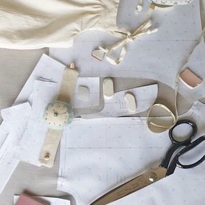 May include: A flat lay of sewing supplies, including a white fabric garment, sewing patterns, scissors, a tape measure, and a small clock. The patterns are labelled "Organic Cotton".