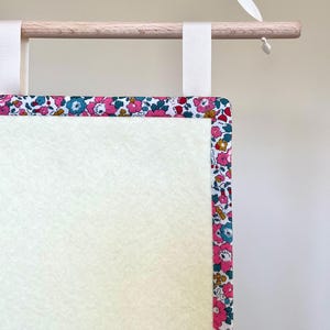 May include: A wall hanging featuring a cream-coloured central panel and a floral border in pink, blue, and yellow. It is suspended from a wooden dowel with white ribbon.