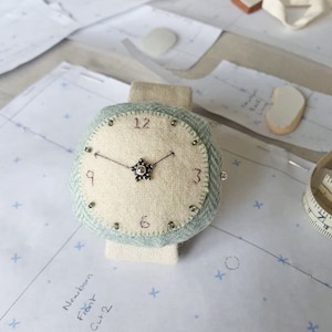 May include: A white and blue fabric watch with a white face and black numbers. The watch is on a white surface with a pattern of small blue stars.