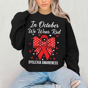 Puede incluir: Sudadera negra con el texto "In October We Wear Red" y "DYSLEXIA AWARENESS". El diseño incluye una cinta roja con lunares blancos y corazones rojos y blancos.