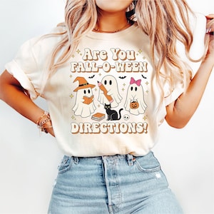 Könnte beinhalten: Cremefarbenes T-Shirt mit einem Retro-Halloween-Design. Die Grafik zeigt drei Geisterfiguren, eine schwarze Katze und den Text "Are You Fall-O-Ween Directions!". Die Geister halten ein Buch, einen Bleistift und einen Kürbis.