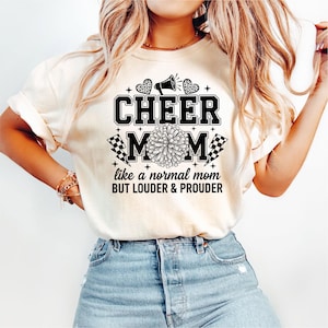 Puede incluir: Camiseta color crema con la frase "CHEER MOM" en negrita y en color negro. El diseño incluye un megáfono, corazones, un pompón y banderas a cuadros. El texto de abajo dice "like a normal mom BUT LOUDER & PROUDER."