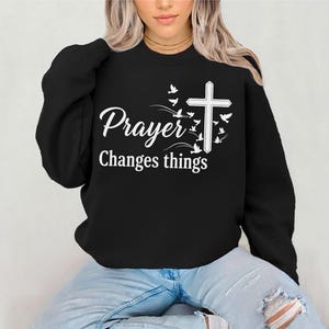 Könnte beinhalten: Schwarzes Sweatshirt mit dem weißen Text "Prayer Changes things". Rechts vom Text sind ein weißes Kreuz und Vögel abgebildet. Das Sweatshirt hat einen Rundhalsausschnitt.