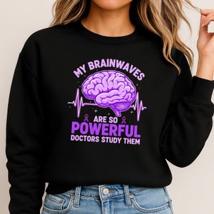 May include: Black sweatshirt with a purple brain graphic and the text "MY BRAINWAVES ARE SO POWERFUL DOCTORS STUDY THEM". The design includes a brain illustration, a heartbeat line, and purple ribbons.