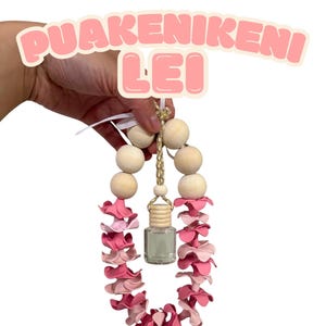 Könnte beinhalten: Ein rosa-weißes Blumen-Lei mit Holzperlen und einer kleinen Glasflasche. Das Lei wird von einer Hand gehalten, und der Text "PUAKENIKENI LEI" befindet sich darüber.